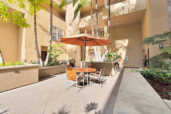Outdoor patio area with table and umbrella, surrounded by palm trees and building walls.