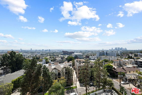 Panoramic city skyline with buildings and trees under a blue sky with clouds.