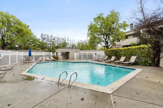 Outdoor swimming pool area with surrounding lounge chairs and a small building in the background.