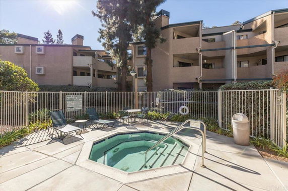 Outdoor area with a Jacuzzi and lounge chairs, surrounded by fencing and apartment buildings.