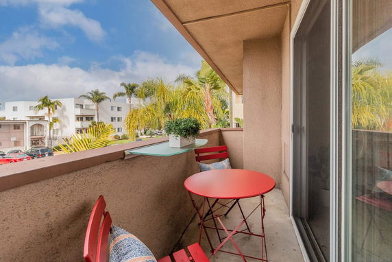 View from a balcony with a red table and chairs, overlooking palm trees and nearby buildings.