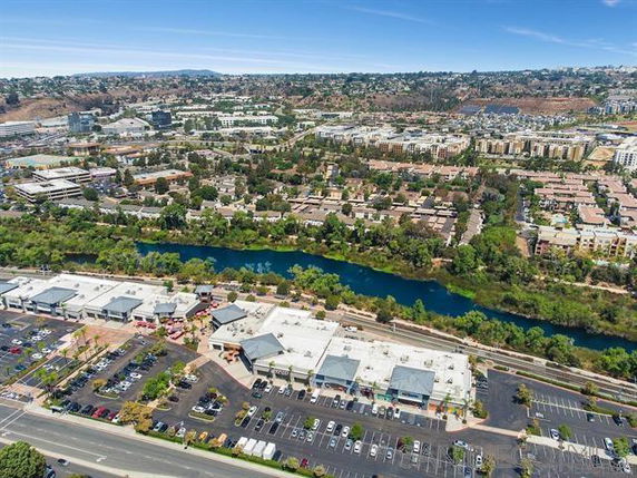 Aerial panoramic view of a shopping area with parking and a river nearby, surrounded by residential and commercial buildings.