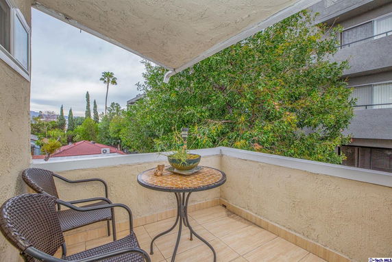 View from a balcony showing a small seating area with a table and two chairs, overlooking nearby buildings and trees.