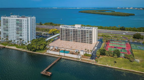 Panoramic view of waterfront buildings with tennis courts and a swimming pool, overlooking a large body of water.