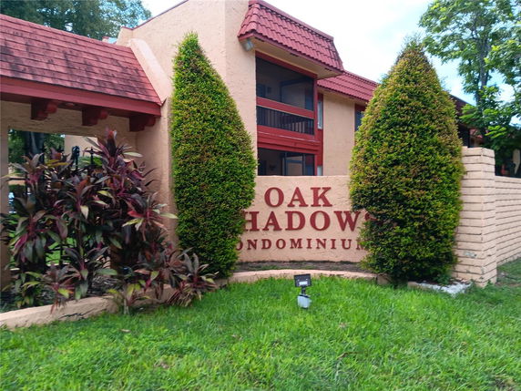 Front view of a condominium building with a brown and red exterior, featuring a sign reading 'Oak Shadow Condominiums' next to trimmed bushes.