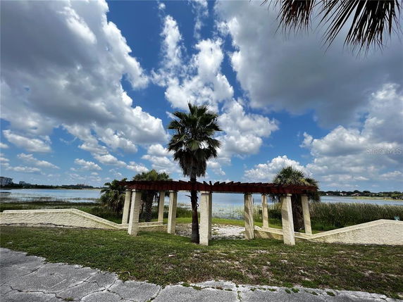 View of a lakeside area with a pergola and palm trees under a partly cloudy sky.