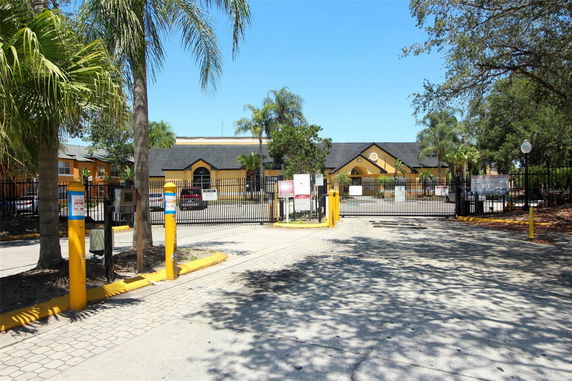 Front view of a gated building entrance with palm trees and a yellow building in the background.