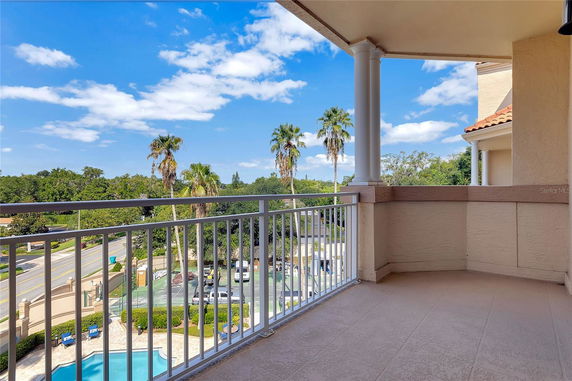 View from a balcony overlooking a pool and tennis court area with palm trees and clear sky.