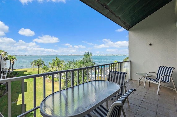 Balcony view overlooking a body of water with distant land and palm trees, featuring patio chairs and a glass table.