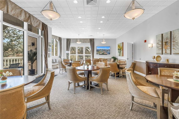 Interior of a clubhouse with round tables and chairs, large windows, and elegant decor.