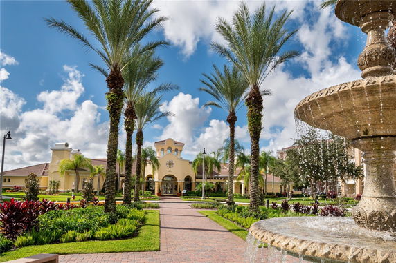Front view of a Mediterranean-style building with arched entrances and decorative elements, framed by tall palm trees and a fountain in the foreground.