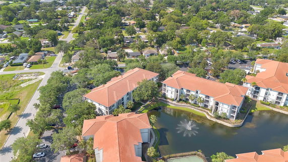 Aerial view of a complex of multi-story residential buildings with red roofs, a pond with a fountain, and surrounding neighborhood.