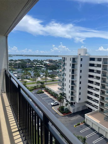 View from a high-rise balcony overlooking buildings, parking area, and distant water body.