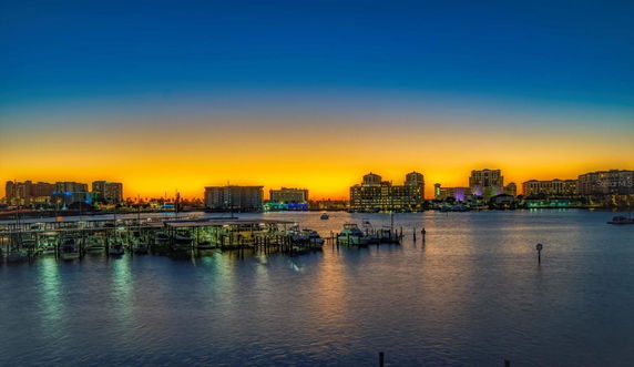 Panoramic view of a waterfront skyline at sunset with illuminated buildings and boats.