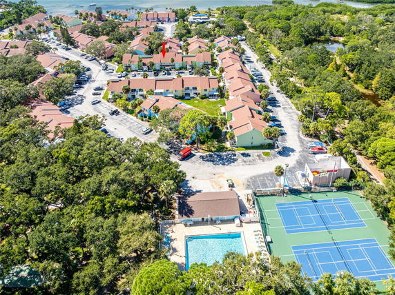 Aerial view of a residential complex with multiple houses, roads, a tennis court, and swimming pool surrounded by trees.