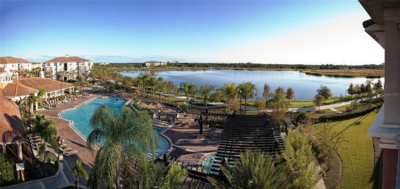 Panoramic view of a large swimming pool area with adjacent buildings and a lake in the background.