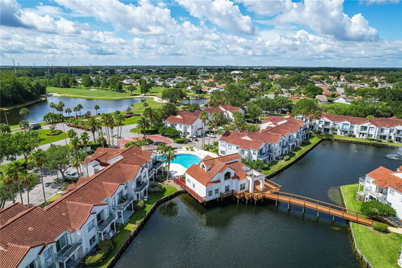 Aerial view of a residential complex with a swimming pool surrounded by buildings with red roofs, and a landscape with water and greenery.