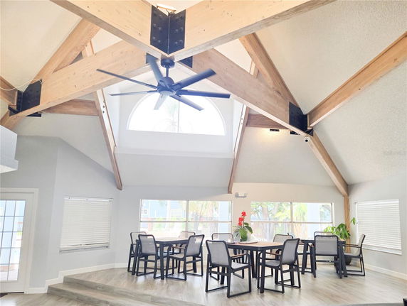 Interior view of a clubhouse with a high ceiling featuring exposed wooden beams, several tables with chairs, and large windows.