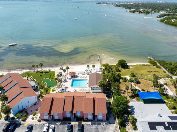 Aerial view overlooking buildings with red roofs, a swimming pool, and a waterfront with a beach and boats.