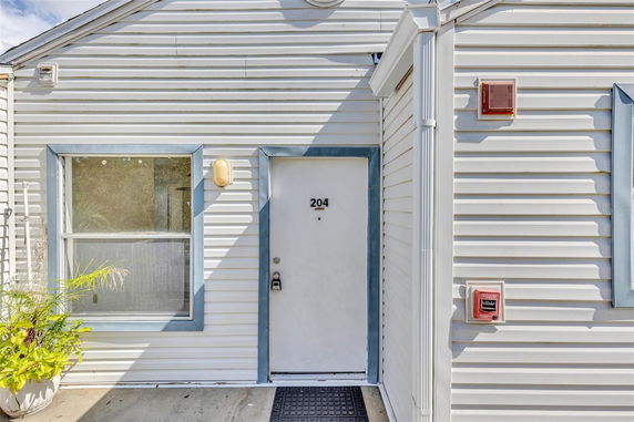 Front view of a house with white siding and a door marked 204, featuring a window and a potted plant.