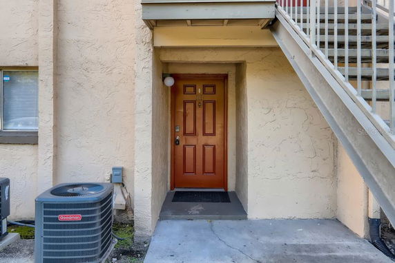 Front entrance of a building with a red door and an air conditioning unit alongside an external staircase.