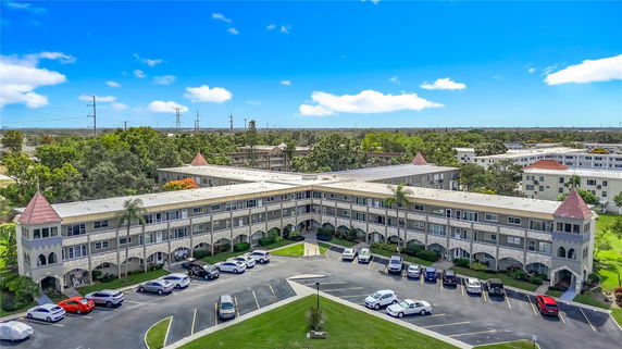 Front view of a multi-story building with a unique architectural design featuring arched elements and corner towers. The structure includes parking space with several cars parked.