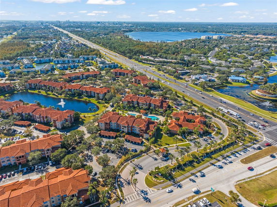 Aerial view of a suburban area with red-roofed buildings, a small pond with a fountain, and surrounding roadways.