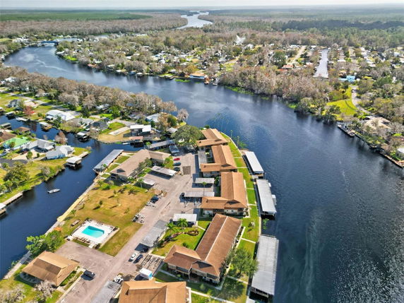 Aerial view of a riverside residential area with multiple buildings and a swimming pool, surrounded by water and trees.
