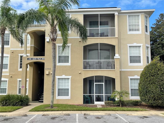Front view of a multi-story apartment building with a beige exterior and white trim, featuring balconies and a central stairway.