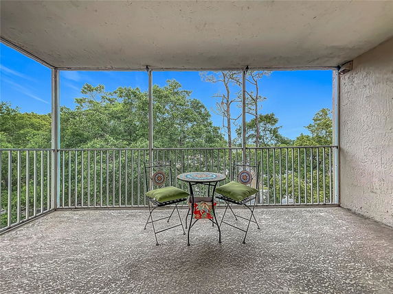 A balcony with a round table and two chairs offers a scenic view of surrounding trees and a clear blue sky.