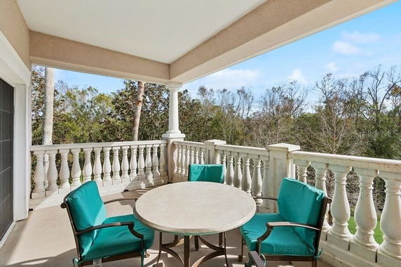 Covered outdoor balcony with a round table and chairs, surrounded by a decorative railing and overlooking trees.