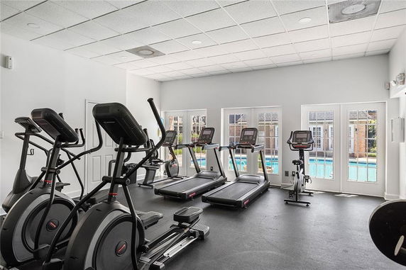 Indoor gym area with exercise equipment overlooking a swimming pool through large windows.