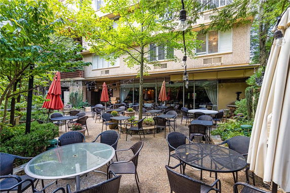 Outdoor seating area with tables and chairs in a garden setting, with a building in the background.