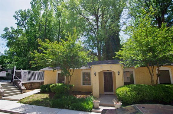 Front view of a single-story building with a yellow exterior and a door in the center, surrounded by trees and shrubs.