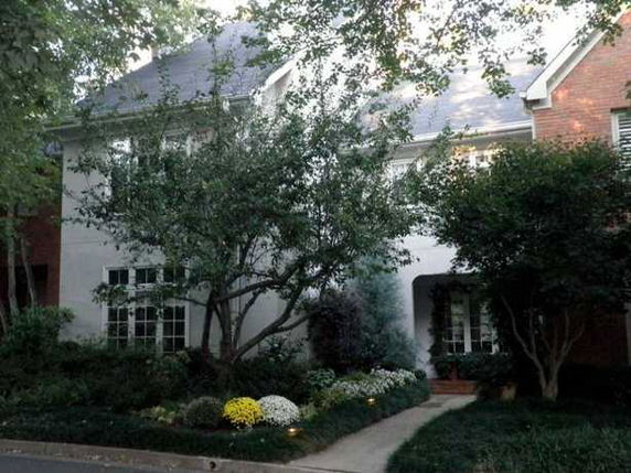 Front view of a two-story house with a sloped roof and large windows, partially obscured by trees and plants.