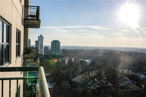 High angle view from a building balcony overlooking a cityscape with trees and distant skyscrapers under a bright sun.