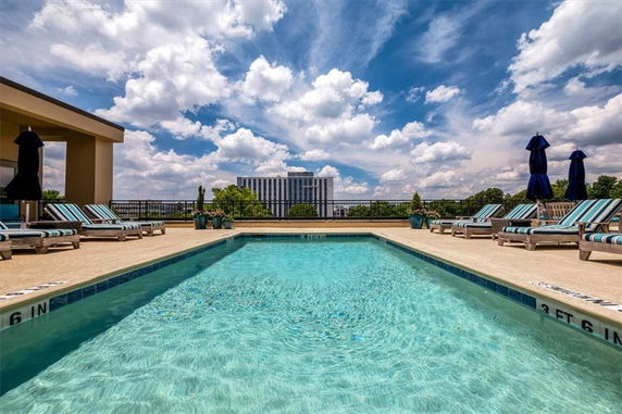 Outdoor swimming pool area with lounge chairs and umbrellas, set against a background of buildings and a blue sky.