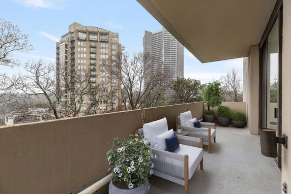 Outdoor balcony with chairs and potted plants overlooking surrounding buildings and trees.