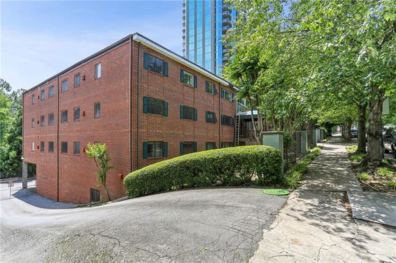 Brick apartment building with multiple windows, situated beside a sidewalk and surrounded by trees.