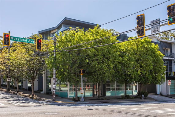 Front view of a modern multi-story building on a street corner with large glass windows and trees in the foreground.
