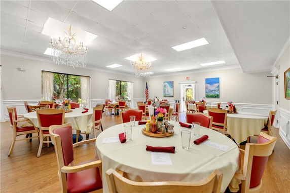 Interior view of a dining area with round tables, red chairs, chandeliers, and large windows in a clubhouse setting.
