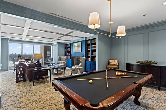 Interior of a room with a billiards table and seating area, featuring bookshelves and large windows.