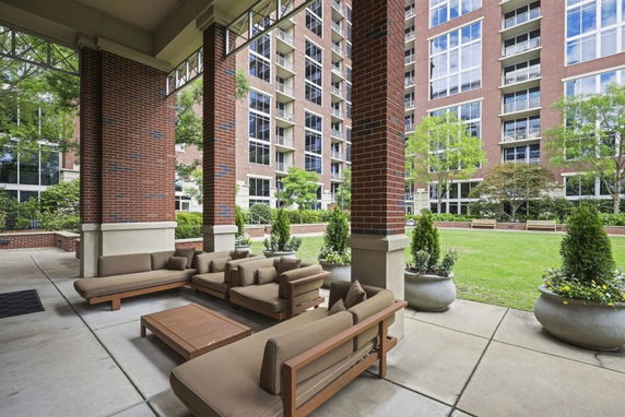 Outdoor seating area with couches and tables under a covered structure, facing a multi-story red brick building.