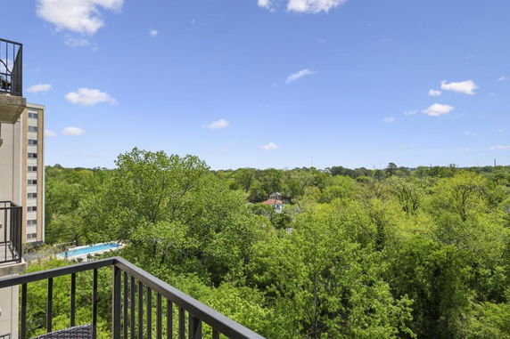 View from a balcony overlooking a forested area with a building and a distant swimming pool visible.