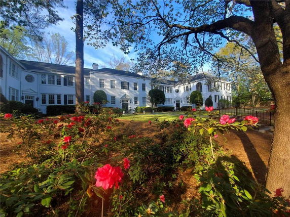 Front view of a large white multi-story building with numerous windows, surrounded by trees and flowers.