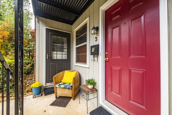 Front view of a porch area with a red door and a black door, featuring a chair and potted plants.
