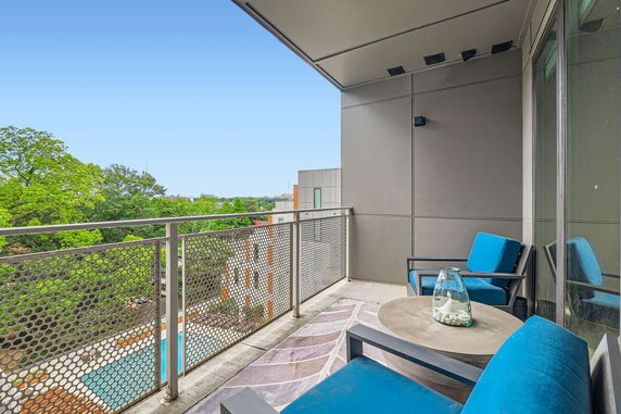 View from a modern balcony with metal railing and seating, overlooking trees and buildings.
