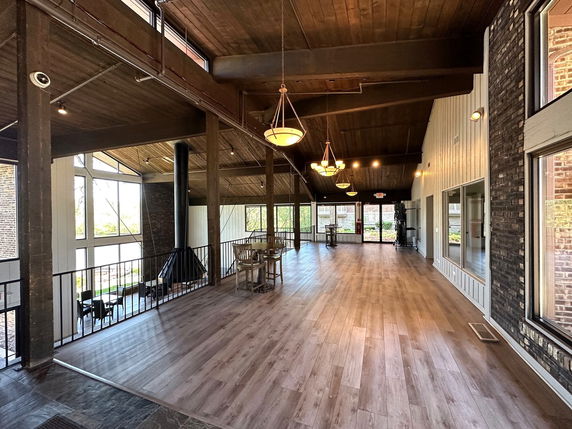 Interior view of a spacious clubhouse with wooden flooring and ceiling, featuring large windows and chandeliers.