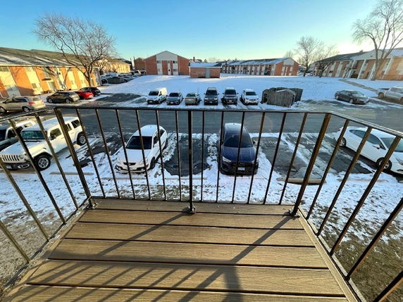 View from a balcony overlooking a snowy parking area with several vehicles and surrounding buildings.