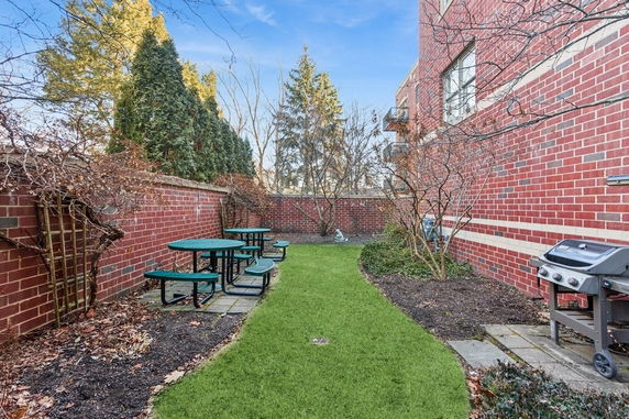 Outdoor patio area with green grass, picnic tables, and a barbecue grill next to a brick building.
